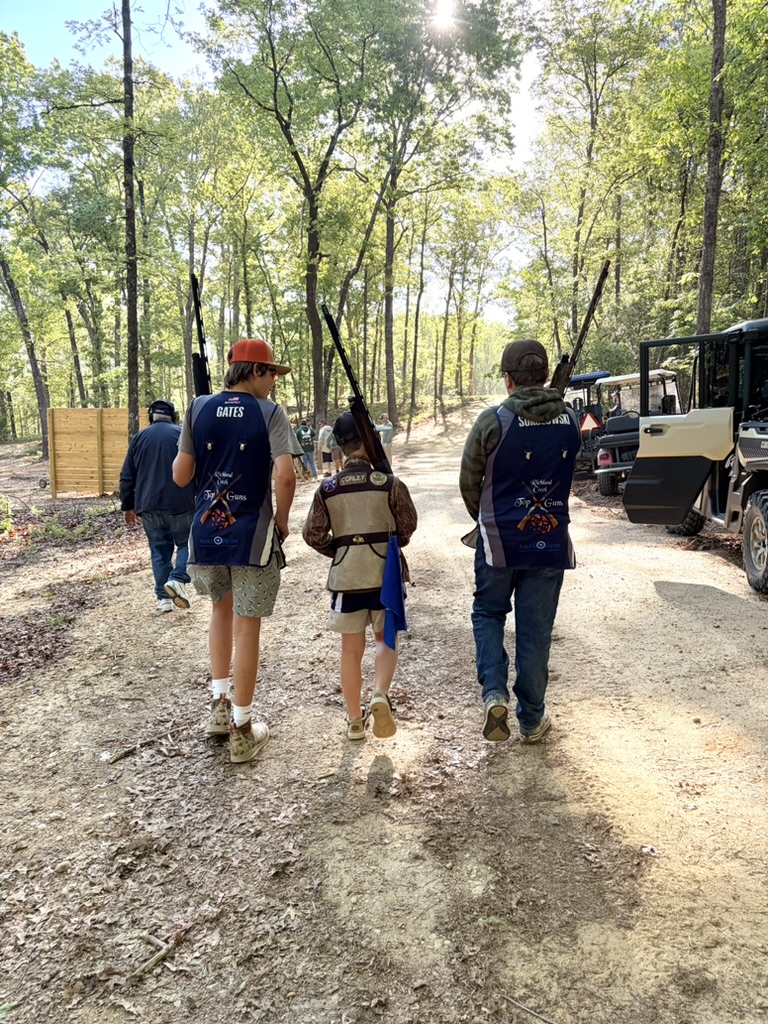 Three Top Guns shooters in team vests walking a wooded course path with shotguns shouldered