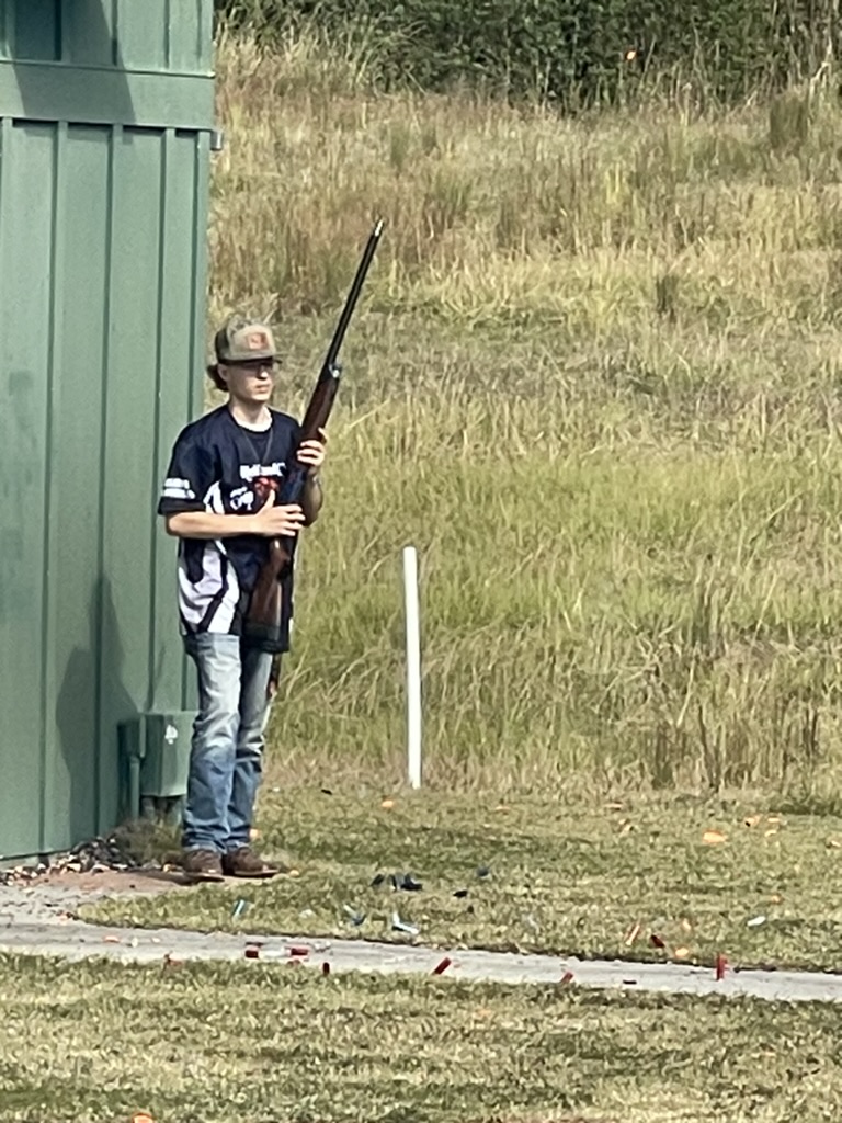 Young shooter in a team jersey waiting with a shotgun beside a green skeet house