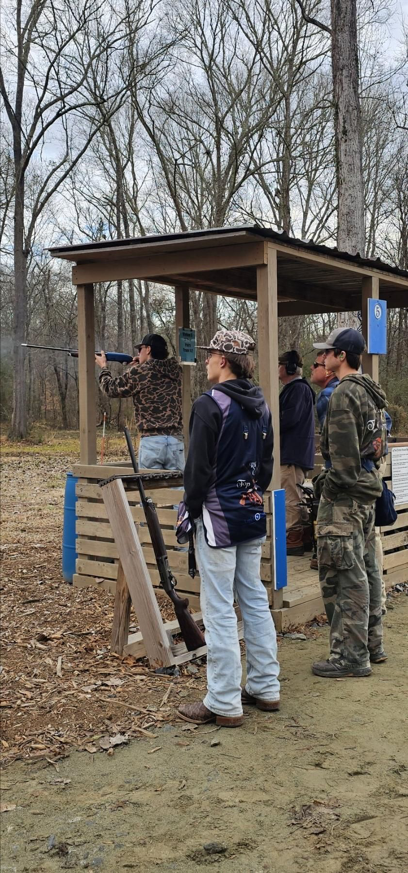 Shooters in team gear waiting behind a covered trap station while a shooter fires