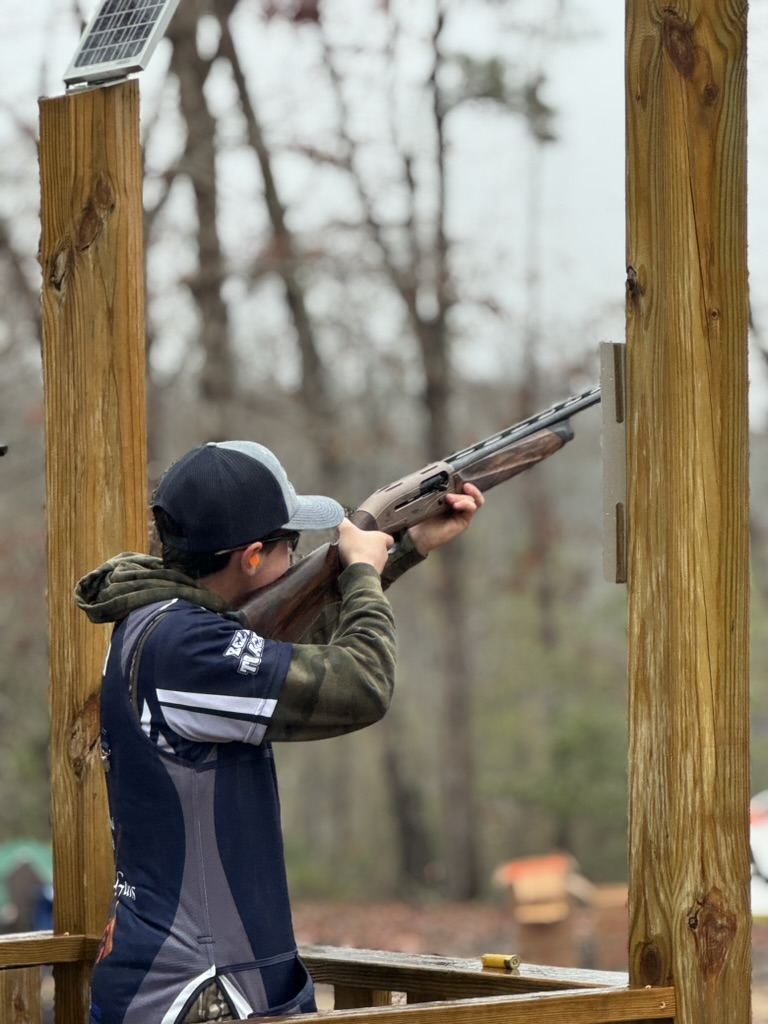 Shooter in a Top Guns vest aiming a shotgun from a covered wooden trap station