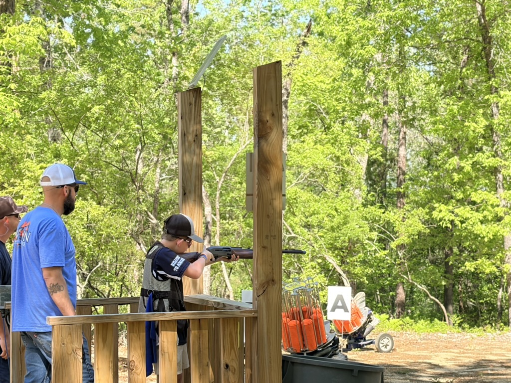 Young shooter aiming from a wooden sporting clays stand while a coach observes