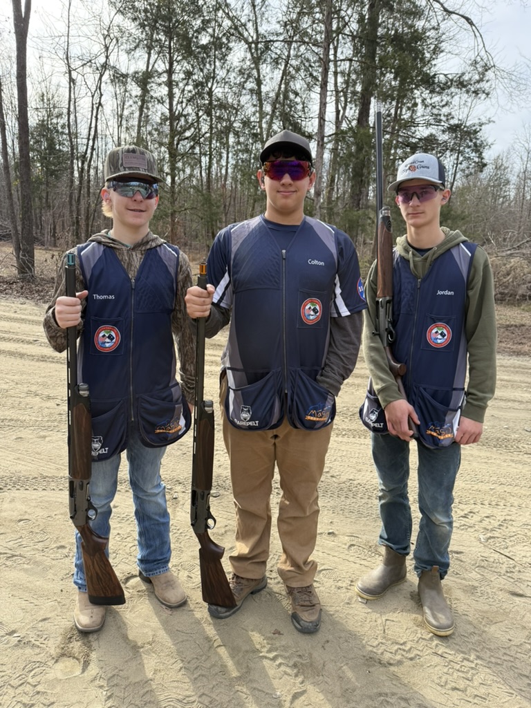 Three Top Guns teammates in team vests holding shotguns on a sandy range road