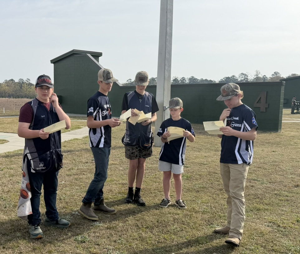 Five Top Guns shooters reading squad sheets on the grass beside trap field four