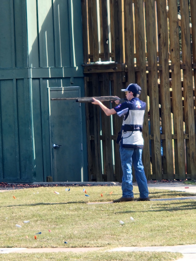 Shooter in a skeet vest firing beside the green high house