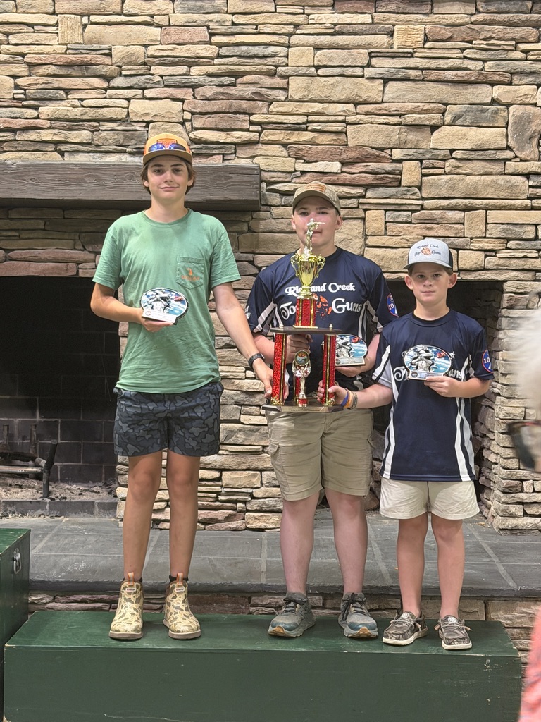 Three young shooters holding trophies and buckles on a podium in front of a stone fireplace