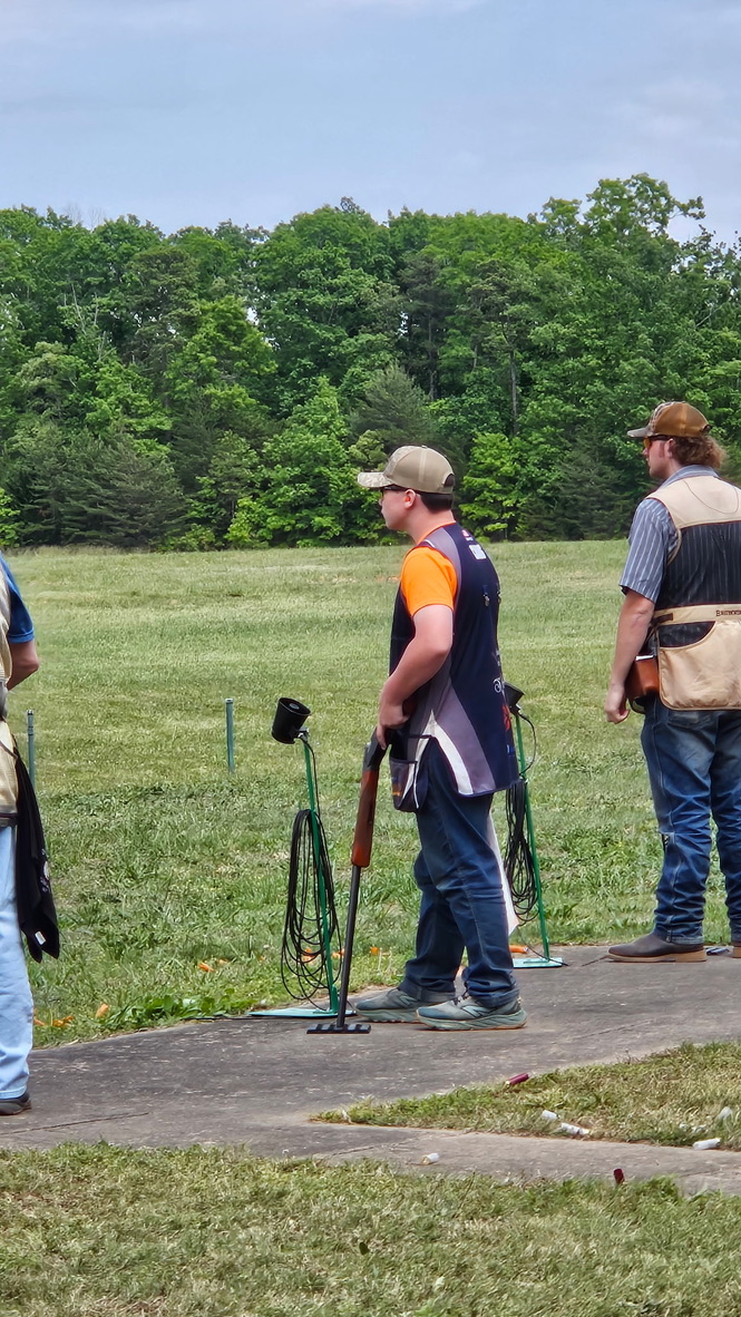 Shooter in an orange shirt and team vest standing at a grass trap line with coaches