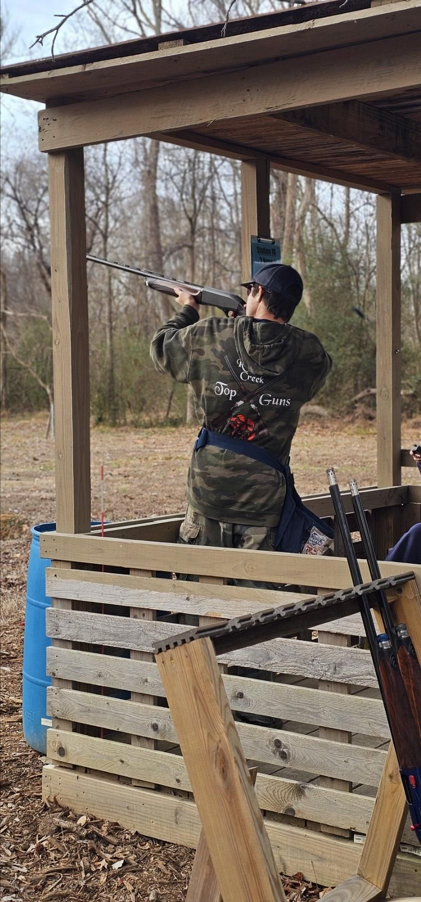 Shooter in a camo Top Guns hoodie firing from a covered sporting clays station