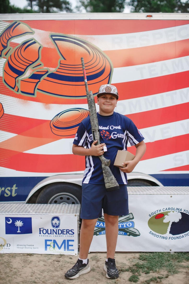 Young shooter in a team jersey holding a shotgun and award box in front of the state shoot trailer