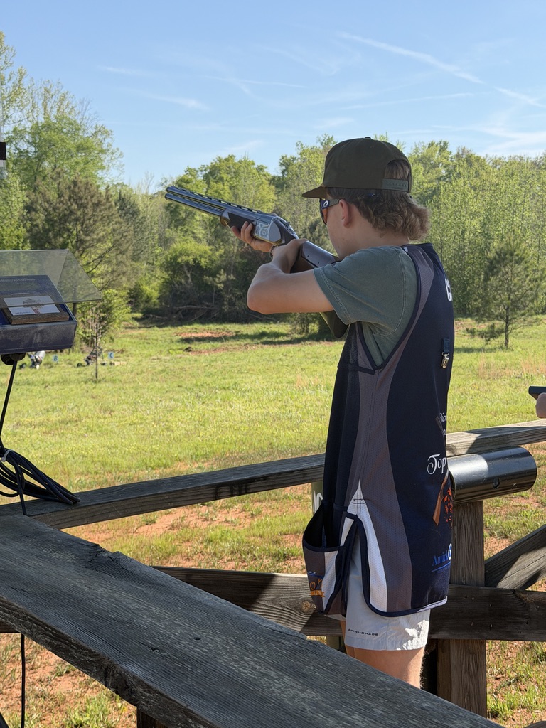 Shooter in a team vest aiming a shotgun over a green field on a sunny day