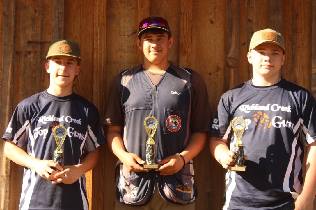 Three Top Guns teammates holding matching trophies in front of a wooden wall