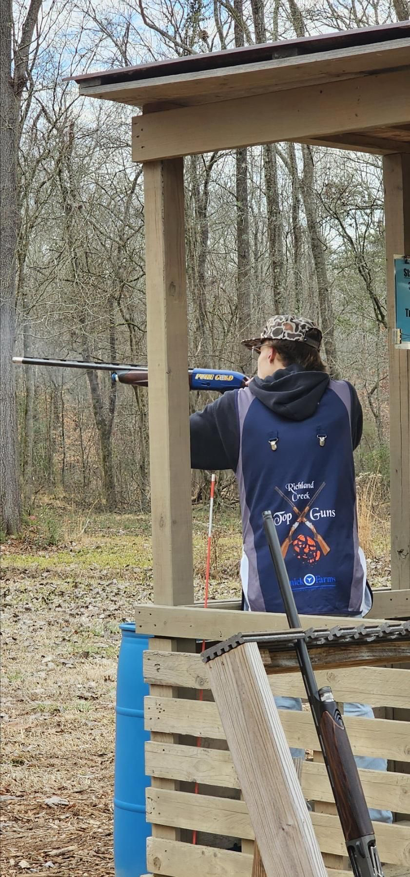 Shooter in a Top Guns vest mounted on a shotgun under a covered wooden station