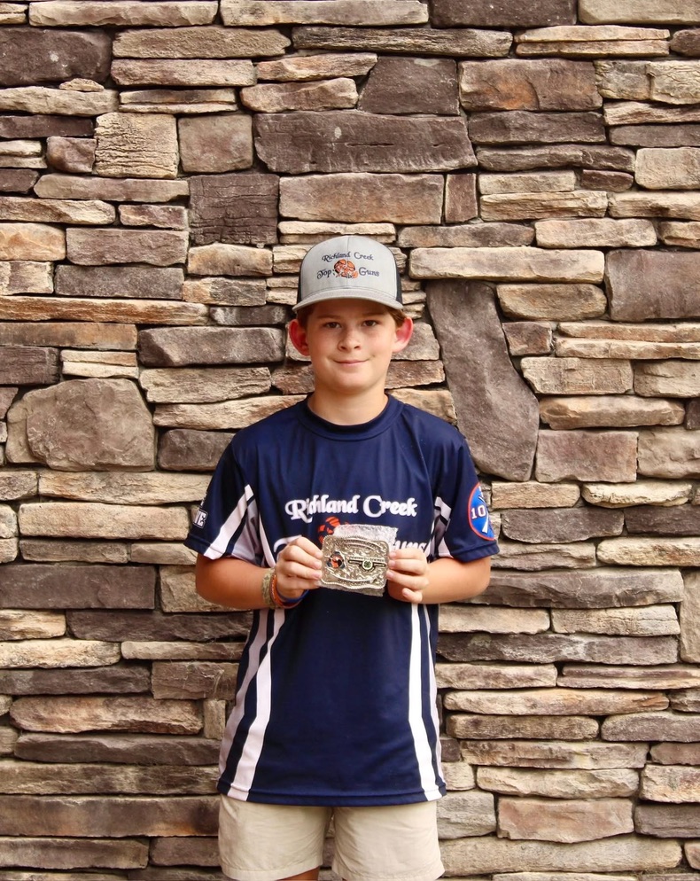 Young shooter in a Richland Creek Top Guns jersey holding a championship belt buckle